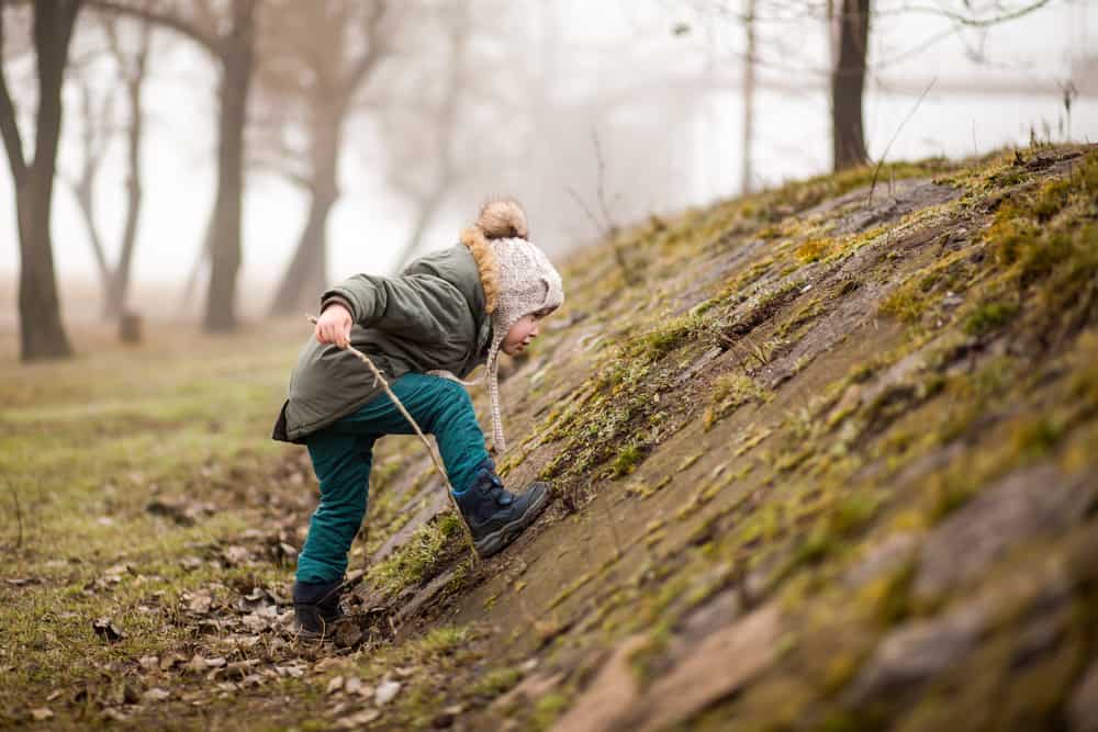 A kid playing on a mossy little hill with sticks.