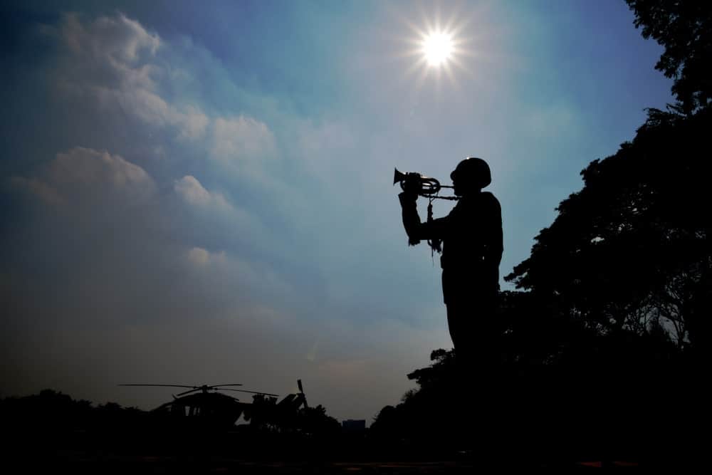 9 Popular Types of Trumpets Explained 16 Silhouette of trees and a soldier blowing a trumpet against a sunny blue sky.