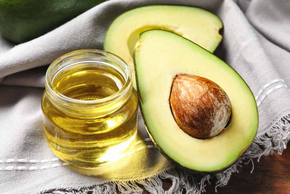 A small jar of avocado oil beside sliced avocados on a gray cloth.