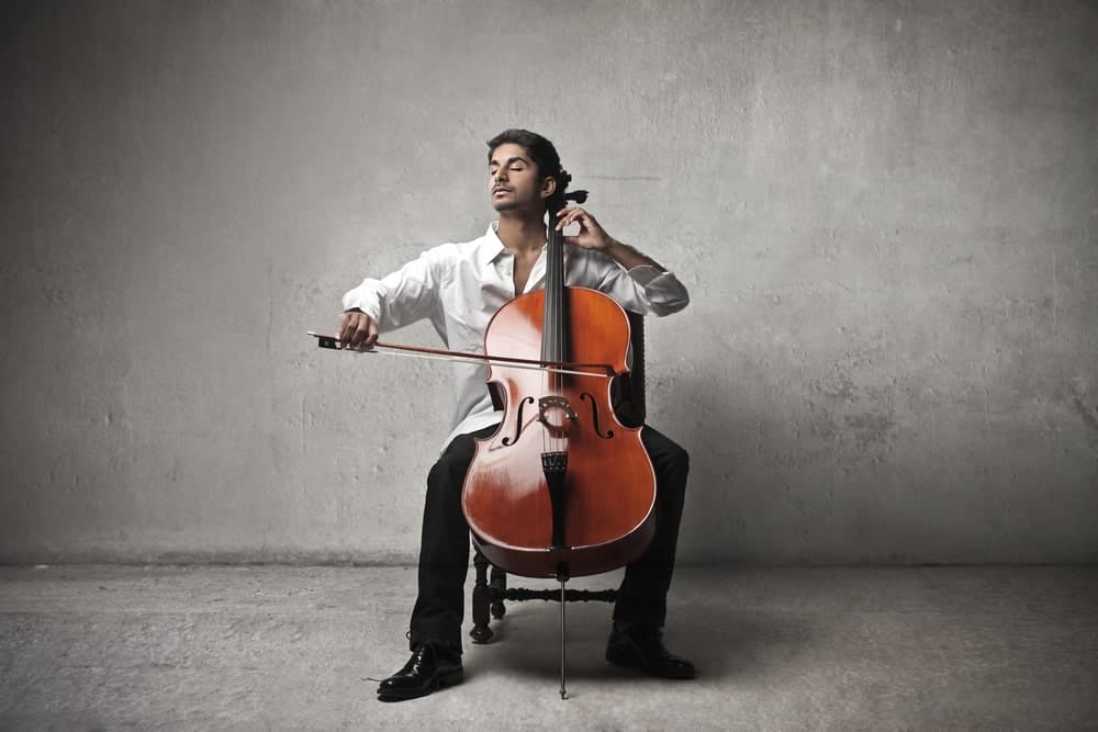 12 Popular Types of Cellos Explained 1 A man sits and closes his eyes playing a cello against a backdrop of gray.