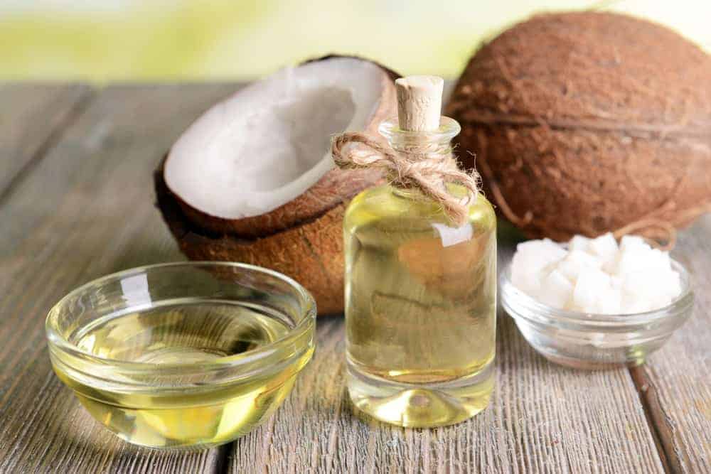 A small bottle and bowl of coconut oil beside coconuts and a bowl of coconut cubes on a wooden surface.