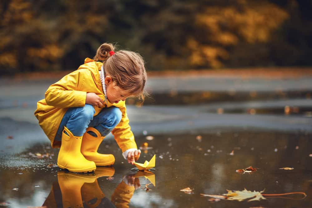 Girl wearing yellow raincoat and boots playing with paper boat on a puddle.