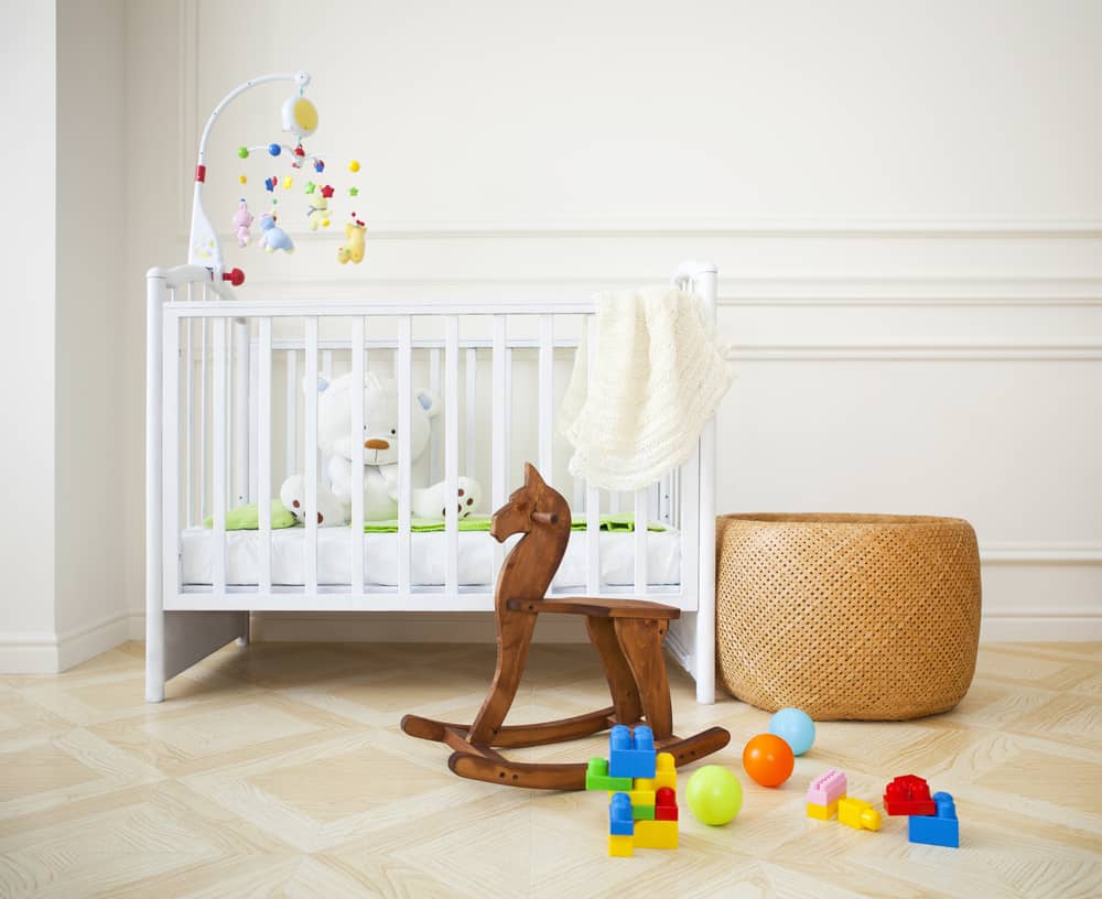 Nursery room with standard crib, wicker ottoman, and a wooden rocking horse.