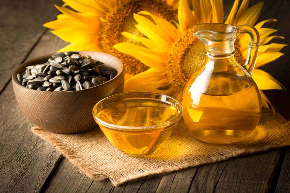 Sunflower oil on a small jar and bowl beside a bowl of sunflower seeds and sunflowers on a rustic background.
