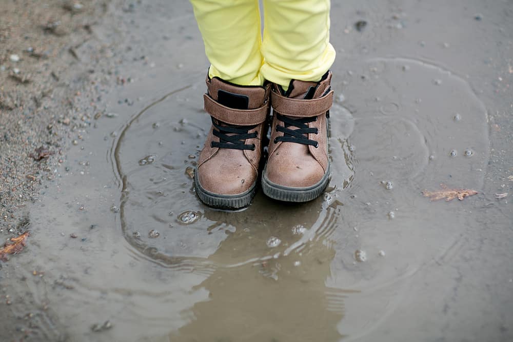 Young boy in work boots standing over a muddy puddle.