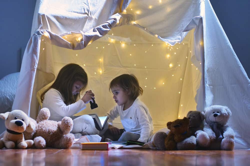 200 Best Indoor Activities for Kids 3 A couple of kids reading in a blanket fort with Christmas lights.