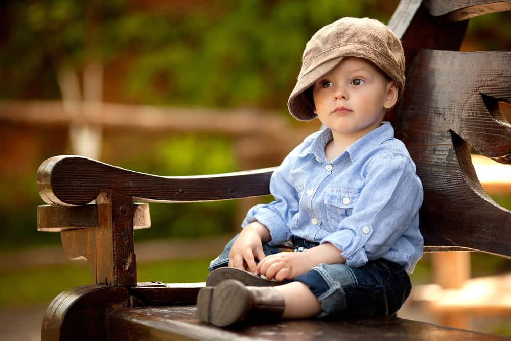 This is a 1-year-old boy on a wooden bench wearing a brown cap.