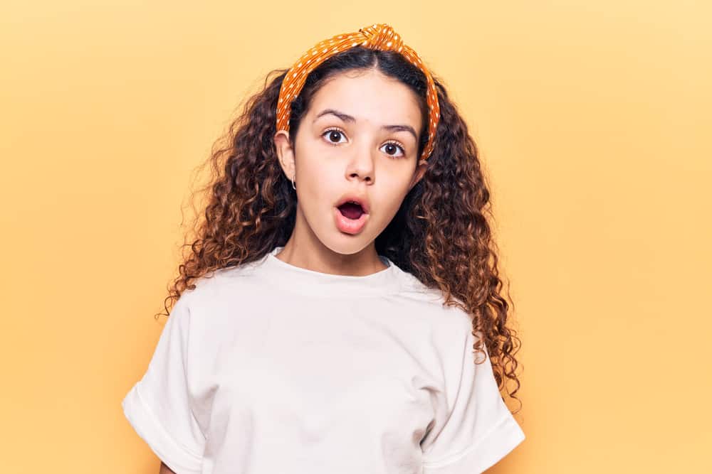A little girl with curly, long hair wearing a dotted headband and white shirt poses with surprised expression against the yellow wall.