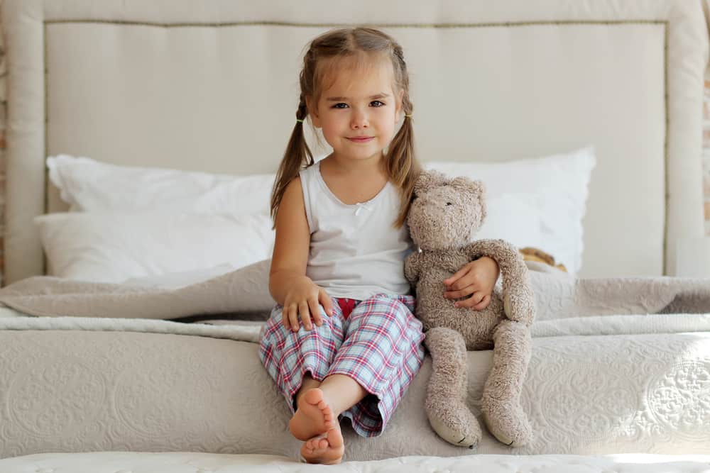 Little girl in a plaid pajama hugging her stuffed toy while sitting on the edge of the bed.