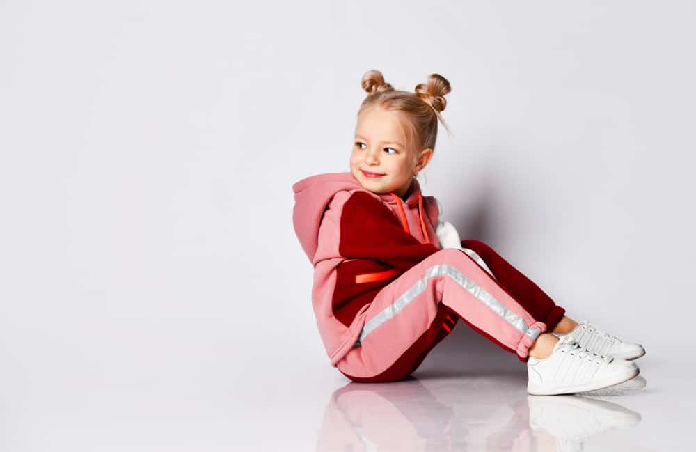 Little girl in pink tracksuit and pigtail buns sitting on the floor.