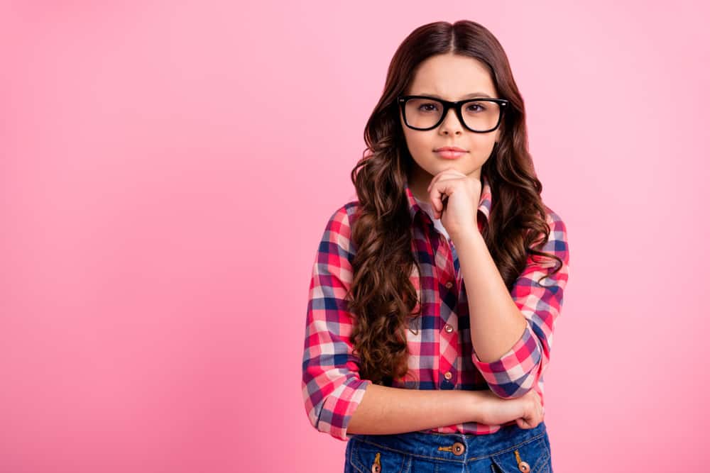 Little girl wearing a plaid long sleeve shirt and glasses posing against the pink wall.