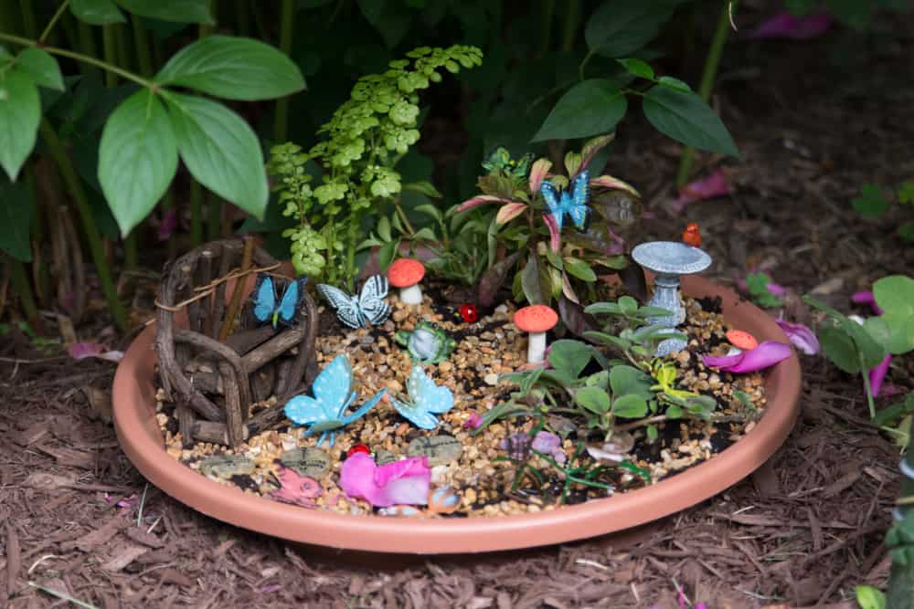 DIY fairy garden in plant saucer filled with butterflies, bird bath, mushrooms, and a driftwood bench.