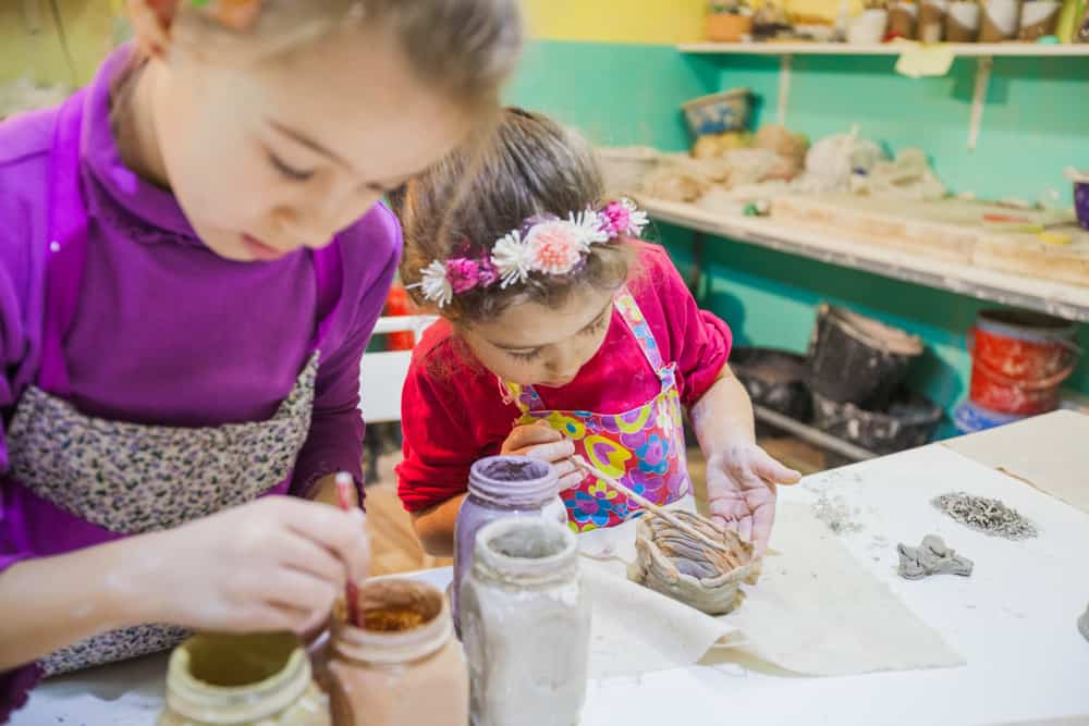 Two little girls painting jars using paint brush and clay.