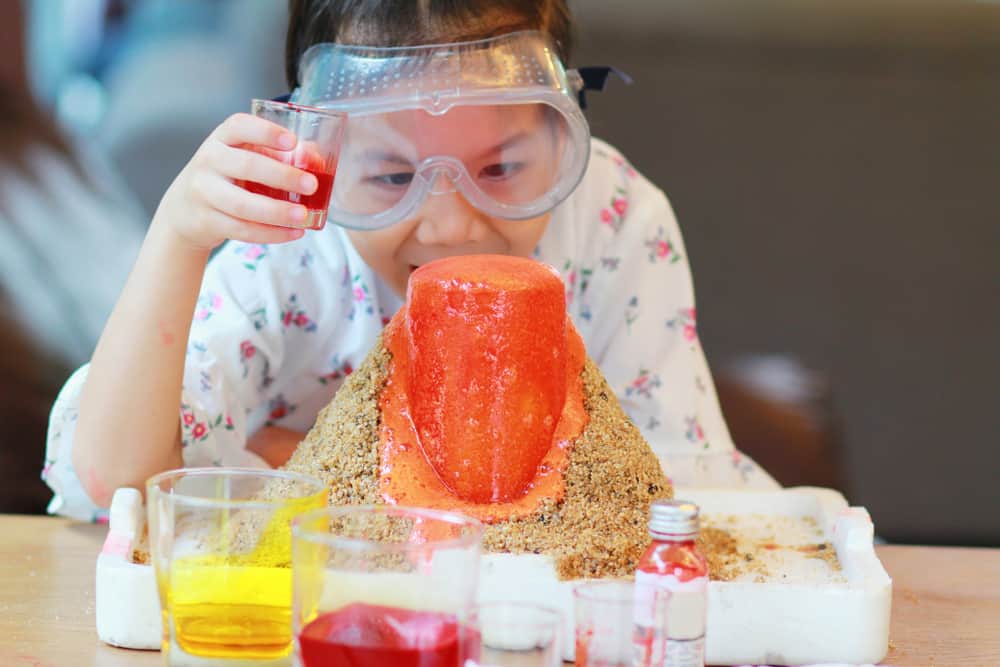 50 Best Science Activities for Kids 7 Little girl in goggles doing a volcano experiment on a wooden table.