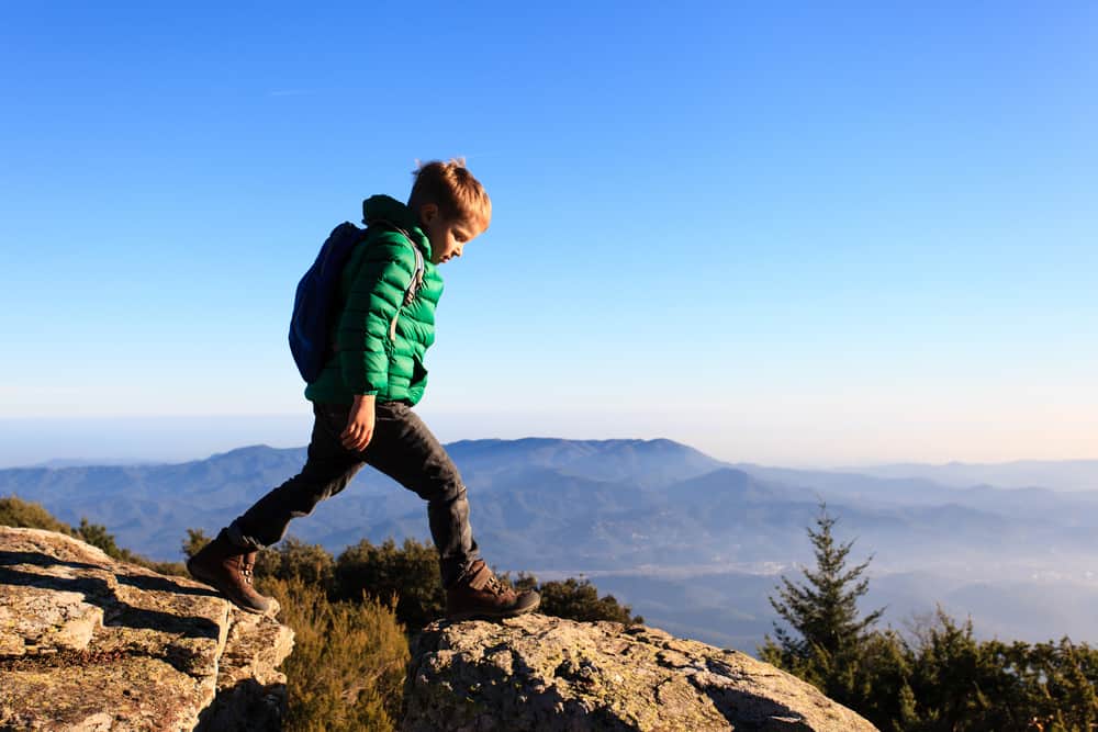 Little boy wearing green winter jacket and backpack hiking along scenic mountains.