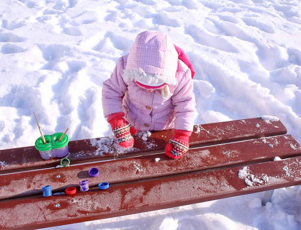 A little girl in pink winter jacket draws gouache on snow.