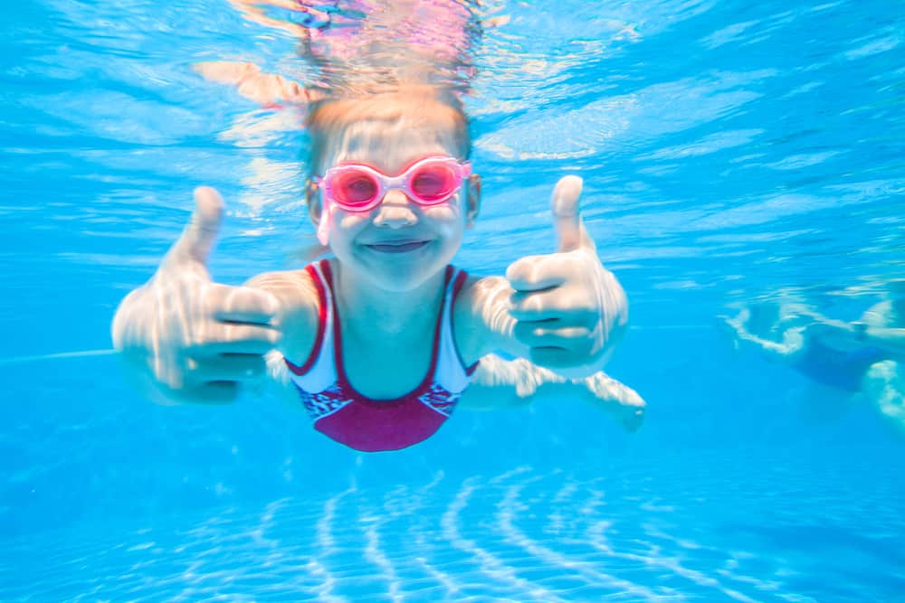 Little girl wearing pink goggles and swimsuit deftly swim underwater in pool.