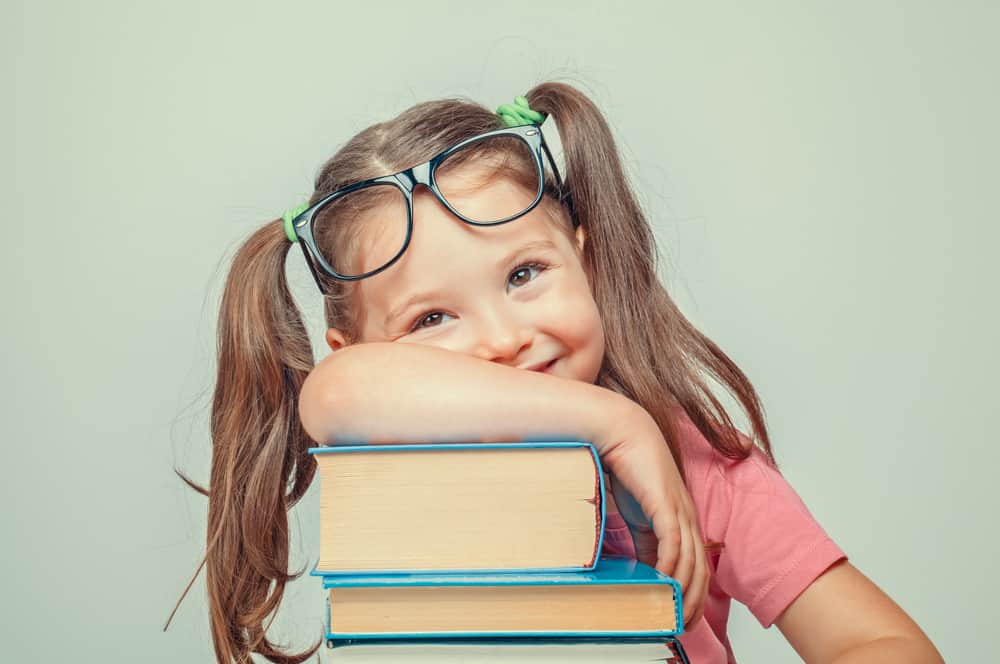 A five-year-old girl with glasses leans on a stack of books.