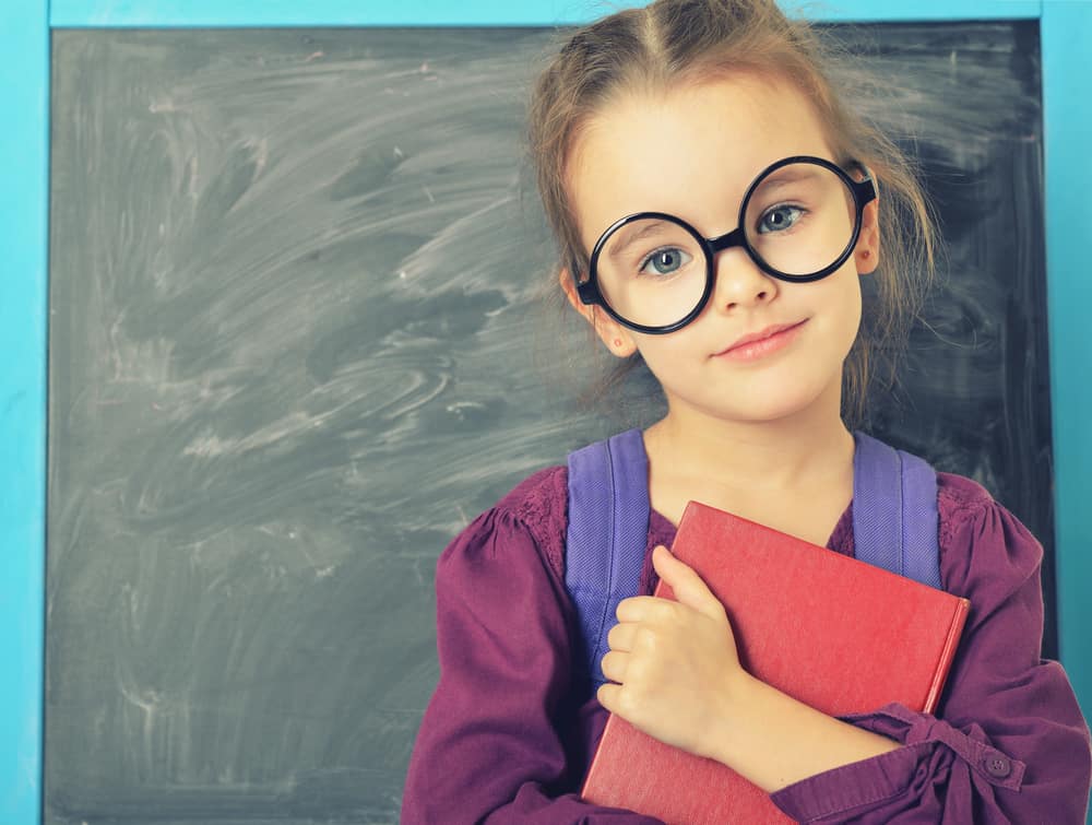 A six-year-old girl with glasses holding a book in front of a blackboard.