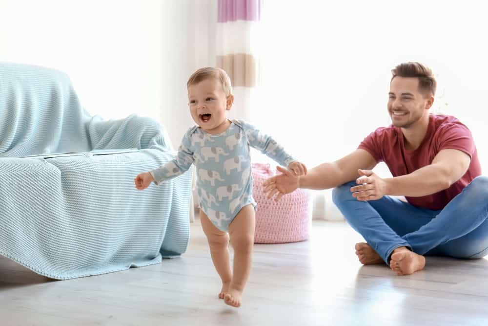 Father guiding her baby girl with her first steps at home.