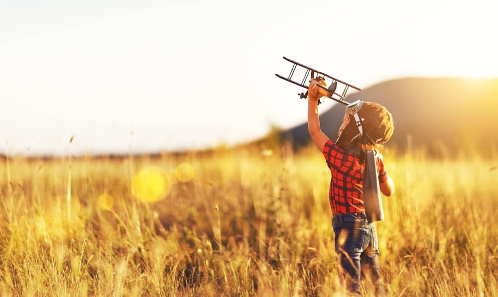 A little boy dressed as a pilot flying an airplane in an open field during sunset.
