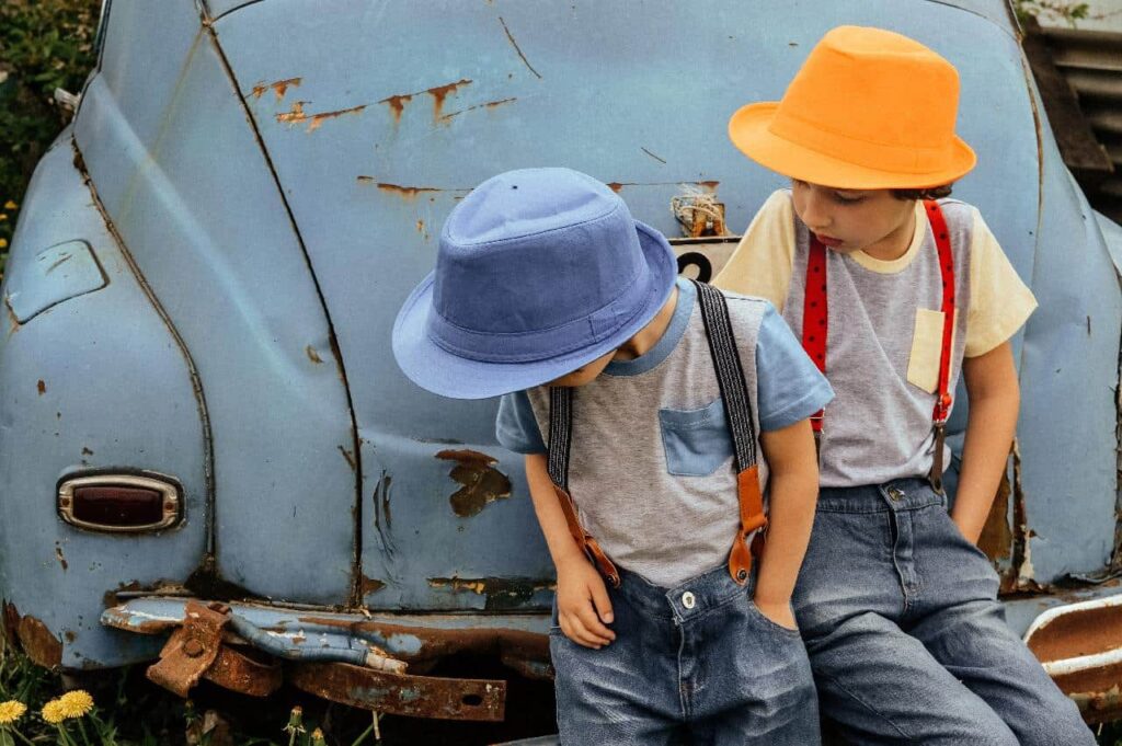 Two little boys in pocketed shirts and hats sitting in front of an old car.