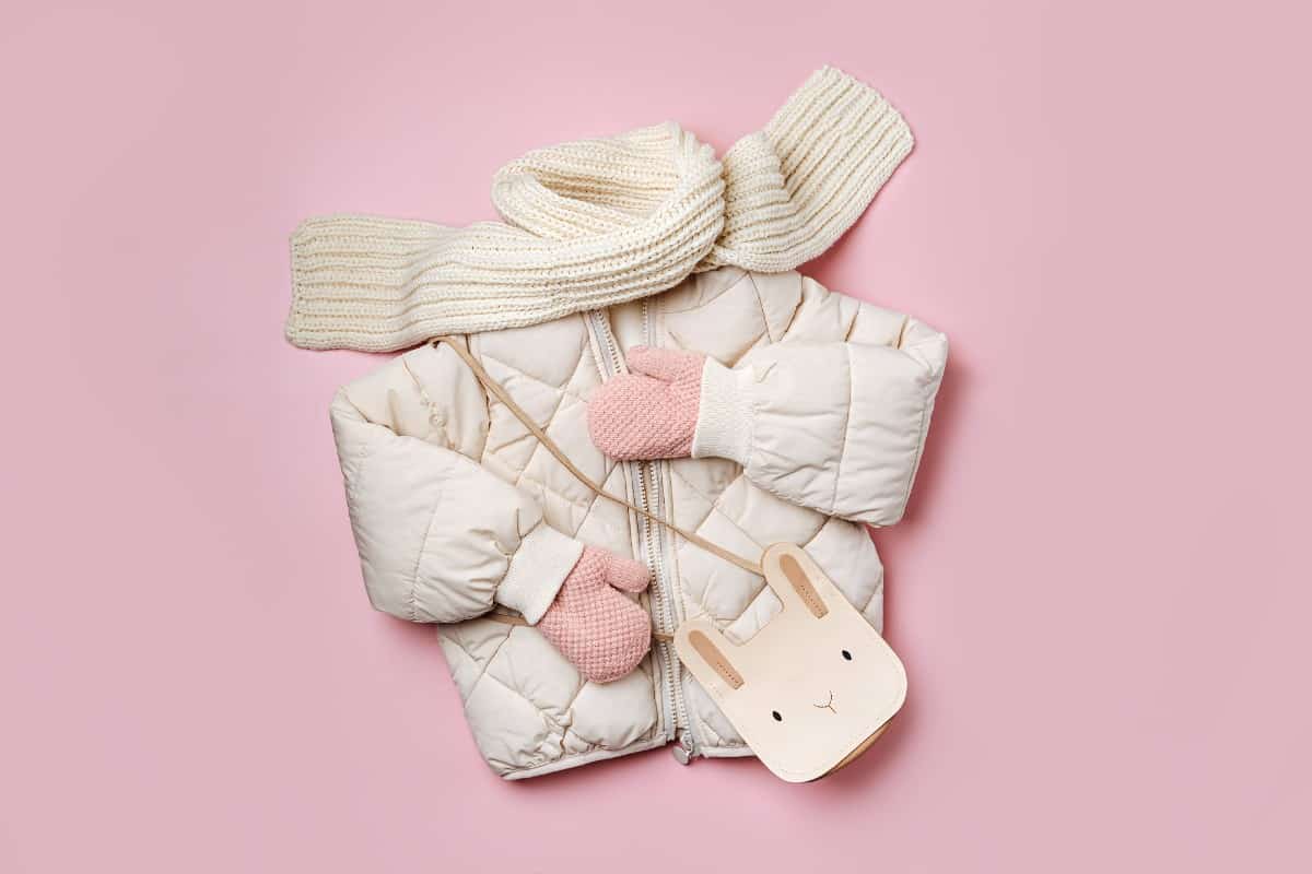 Kids puffer jacket with scarf and gloves against pink background.